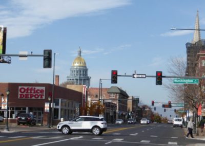 A white car drives along a street in Denver, CO, with urban landscape and clear skies