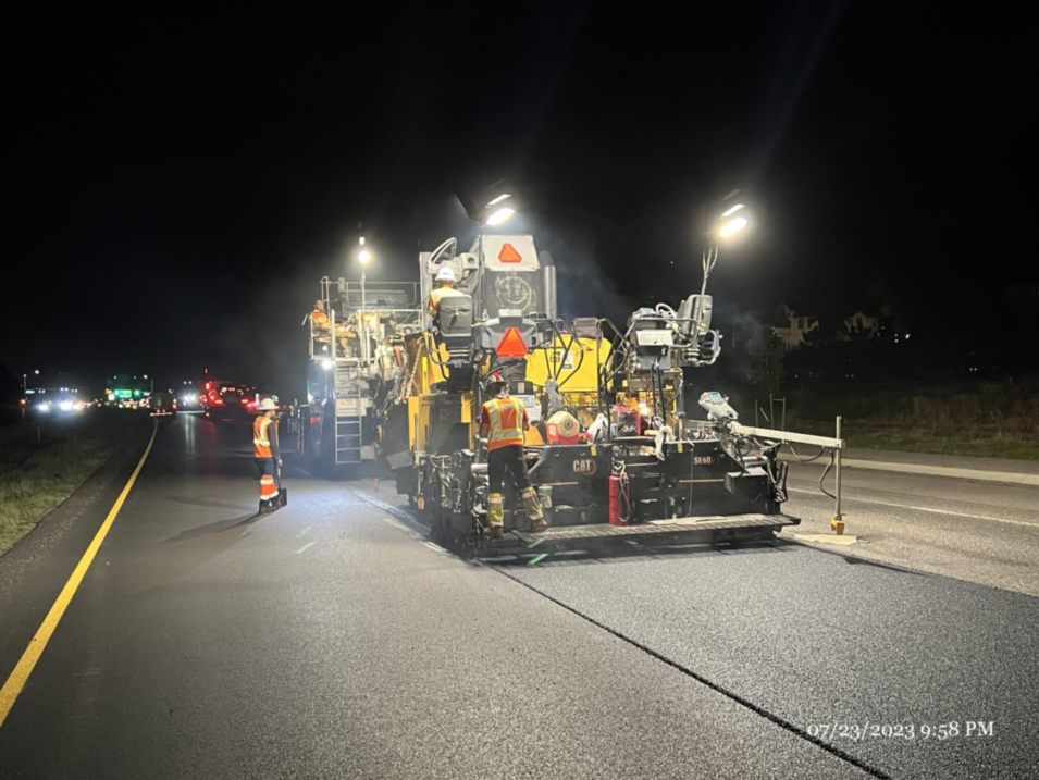 Men work on a road at night in Denver, CO, illuminated by streetlights and construction equipment